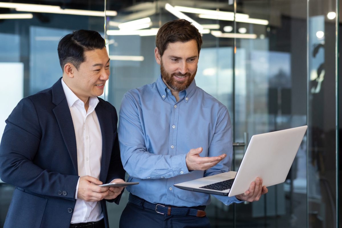 Two men in business attire stand in an office; one holds a laptop and gestures to the screen while the other holds a tablet and smiles.