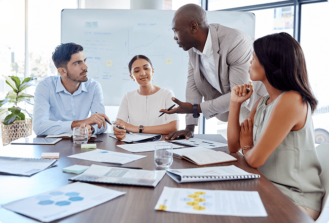 Four professionals sit at a conference table with documents while one person stands and speaks, suggesting a business meeting or discussion in an office setting.
