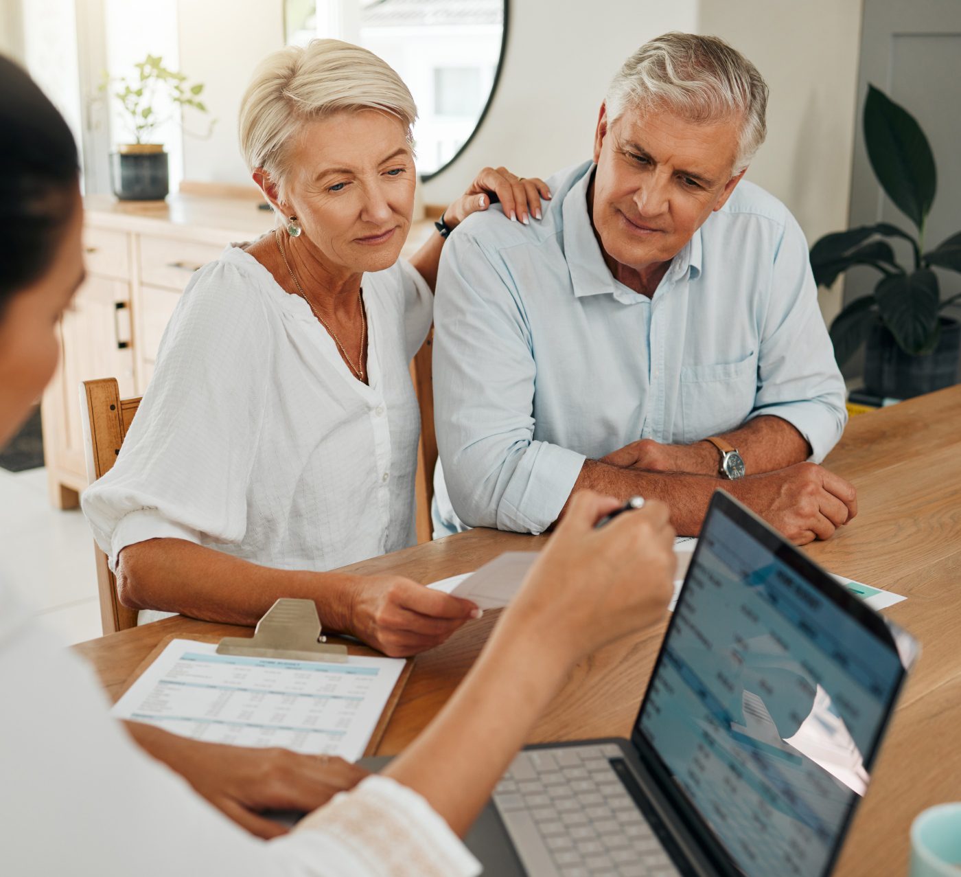 An older couple sits at a table reviewing documents with a professional, who gestures to financial charts on a laptop screen.