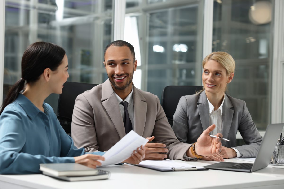 Three business professionals in formal attire sit at a table in an office, engaged in discussion with documents and a laptop in front of them.
