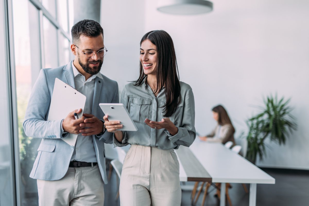 Two people in business attire stand and look at a tablet together, smiling, while another person works at a desk in the background in a modern office.