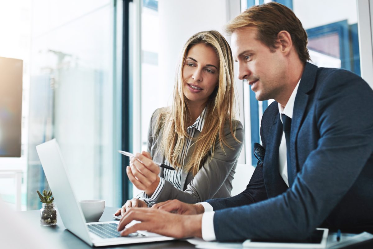 Two business professionals sit at a desk, looking at a laptop screen; the woman points at the screen while the man types on the keyboard.