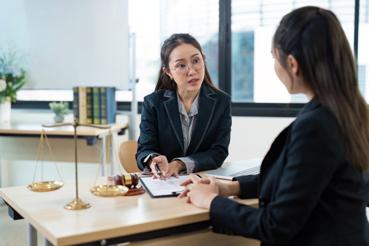 Two women in business attire sit at a desk discussing documents. Legal scales, a gavel, and books are on the table, suggesting a legal or consultation setting.